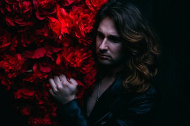 acımasız adam uzun saçlı çıplak bir vücut üzerinde deri ceketli kırmızı çiçek çiçekli dekor yakın duruyor. üzgün ve umutsuz bir yalnız adam