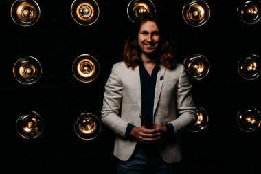 beyaz bir ceket ve siyah tişört, uzun saçlı bir adam parlak fenerler, safites fonunda duruyor. Sahne Alanı'nda. performans. gülümsüyor