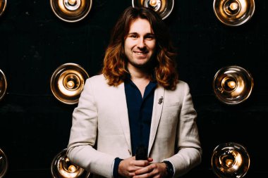 beyaz bir ceket ve siyah tişört, uzun saçlı bir adam parlak fenerler, safites fonunda duruyor. Sahne Alanı'nda. performans. gülümsüyor