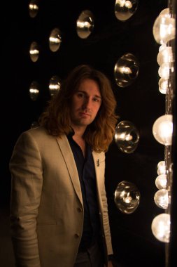 beyaz bir ceket ve siyah tişört, uzun saçlı bir adam parlak fenerler, safites fonunda duruyor. Sahne Alanı'nda. performans