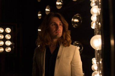 beyaz bir ceket ve siyah tişört, uzun saçlı bir adam parlak fenerler, safites fonunda duruyor. Sahne Alanı'nda. performans