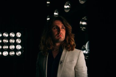 beyaz bir ceket ve siyah tişört, uzun saçlı bir adam parlak fenerler, safites fonunda duruyor. Sahne Alanı'nda. performans