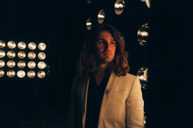 beyaz bir ceket ve siyah tişört, uzun saçlı bir adam parlak fenerler, safites fonunda duruyor. Sahne Alanı'nda. performans