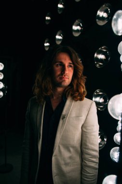 beyaz bir ceket ve siyah tişört, uzun saçlı bir adam parlak fenerler, safites fonunda duruyor. Sahne Alanı'nda. performans