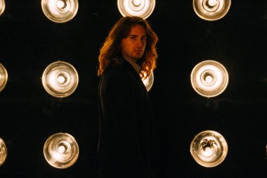 portre parlak fenerler, safites fonunda bir siyah ceket ve siyah tişört, uzun saçlı bir adam duruyor. Sahne Alanı'nda. Performance.Turn çevresinde