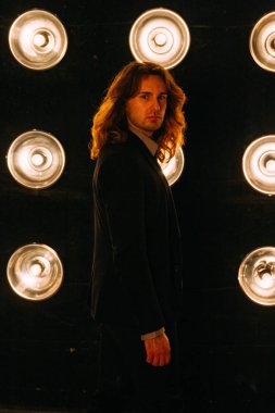 portre parlak fenerler, safites fonunda bir siyah ceket ve siyah tişört, uzun saçlı bir adam duruyor. Sahne Alanı'nda. Performance.Turn çevresinde