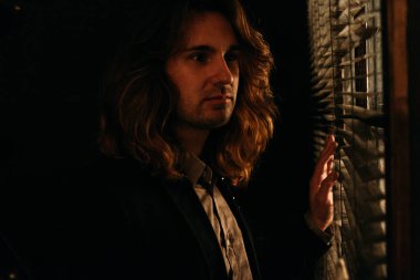 uzun saçlı bir adam güneşlikler loblar, siyah ceketli açılarak pencerenin duruyor. Sokakta insanlar üzerinde casus bakmak gündoğumu. pencereden dışarı bakıp