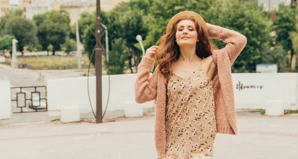 Sonriente chica feliz en vestido y sombrero en una calle de la ciudad y mirando a la cámara. en un sombrero, un vestido en una flor y una chaqueta de colores cálidos —  Fotos de Stock