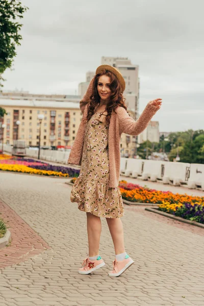 Sorrindo menina feliz em vestido e chapéu em uma rua da cidade e olhando para a câmera. em um chapéu, um vestido em uma flor e uma jaqueta de cores quentes — Fotografia de Stock