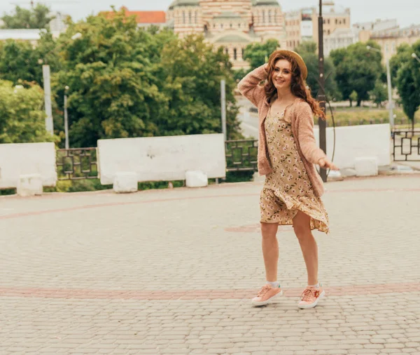 Sorrindo menina feliz em vestido e chapéu em uma rua da cidade e olhando para a câmera. em um chapéu, um vestido em uma flor e uma jaqueta de cores quentes — Fotografia de Stock