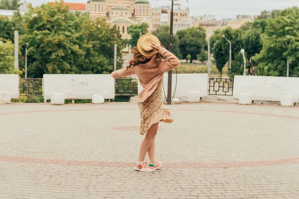 Sonriente chica feliz en vestido y sombrero en una calle de la ciudad y mirando a la cámara. en un sombrero, un vestido en una flor y una chaqueta de colores cálidos — Foto de Stock