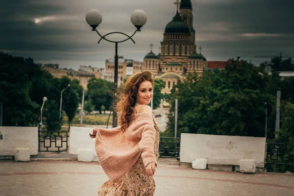 Souriant fille heureuse en robe et chapeau dans une rue de la ville et regardant la caméra. dans un chapeau, une robe dans une fleur et une veste de couleurs chaudes — Photo