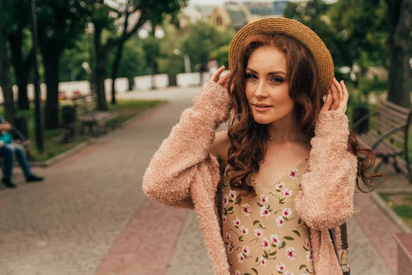 Sonriente chica feliz en vestido y sombrero en una calle de la ciudad y mirando a la cámara. en un sombrero, un vestido en una flor y una chaqueta de colores cálidos —  Fotos de Stock