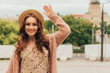 Mutlu kız elbise ve şapka bir şehir sokak ve kamera bakarak gülümsüyordu. bir şapka, bir elbise bir çiçek ve sıcak renk bir ceket. o tebrik için elini kaldırdı. Selamlar, Toplantı sevinci