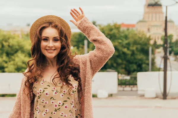 Sorrindo menina feliz em vestido e chapéu em uma rua da cidade e olhando para a câmera. em um chapéu, um vestido em uma flor e uma jaqueta de cores quentes. Ela levantou a mão para cumprimentar. saudação, alegria do encontro — Fotografia de Stock