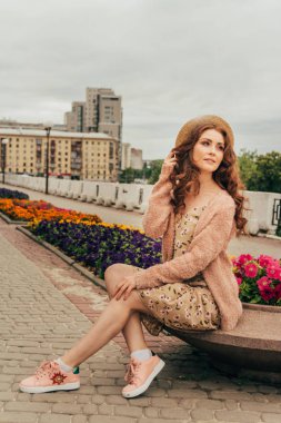 şapkalı, güzel bir kız saç rüzgardan tutar. şehir etrafında yürümek. Kızıl saçlı bir kız portresi. Çiçek, bej kazak ve hasır şapka bej elbise. aşk ve cinsellik. bir çiçek yatak çiçeklerle oturur