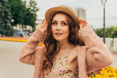 şapkalı, güzel bir kız saç rüzgardan tutar. şehir etrafında yürümek. Kızıl saçlı bir kız portresi. Çiçek, bej kazak ve hasır şapka bej elbise. aşk ve cinsellik. bir çiçek yatak çiçeklerle oturur