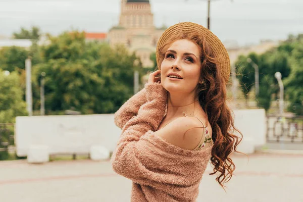 a beautiful girl in a hat, keeps the hair from the wind. walk around the city. portrait of a red-haired girl. Beige dress in flower, beige sweater and straw hat. romance and sexuality