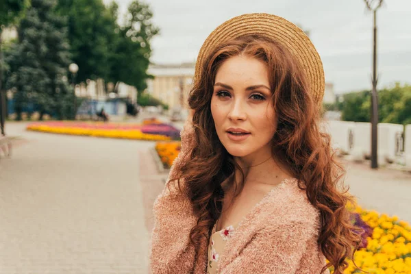 a beautiful girl in a hat, keeps the hair from the wind. walk around the city. portrait of a red-haired girl. Beige dress in flower, beige sweater and straw hat. romance and sexuality. sits near a flower bed with flowers