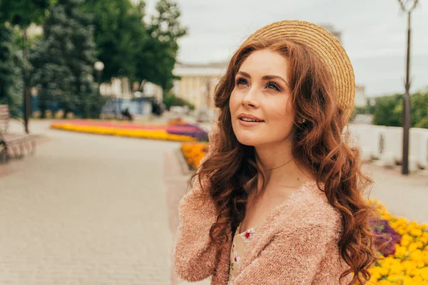 a beautiful girl in a hat, keeps the hair from the wind. walk around the city. portrait of a red-haired girl. Beige dress in flower, beige sweater and straw hat. romance and sexuality. sits near a flower bed with flowers