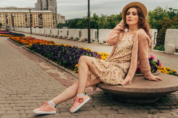 a beautiful girl in a hat, keeps the hair from the wind. walk around the city. portrait of a red-haired girl. Beige dress in flower, beige sweater and straw hat. romance and sexuality. sits near a flower bed with flowers. poses