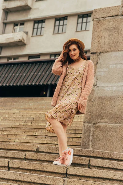 a beautiful girl in a hat, keeps the hair from the wind. stands on the scaffolds. walk around the city. portrait of a red-haired girl. Beige dress in flower, beige sweater and straw hat. romance and sexuality