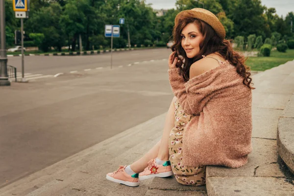 a beautiful girl in a hat, keeps the hair from the wind. sits on scraps. walk around the city. portrait of a red-haired girl. Beige dress in flower, beige sweater and straw hat. romance and sexuality. flirting