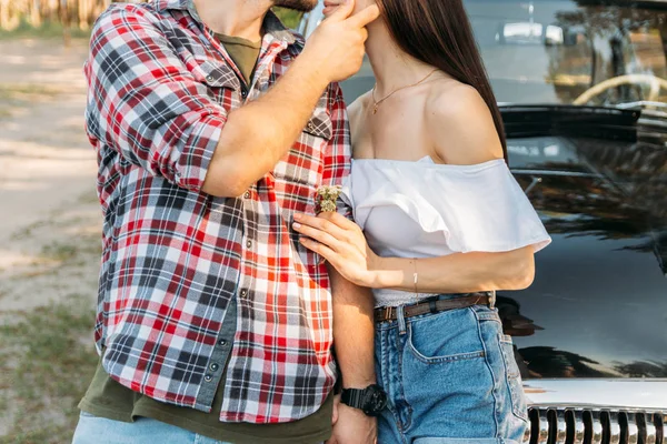 Love and affection between a young couple at the park. a guy in a plaid plane and jeans, a girl in shorts and a white jacket. They get together in the forest for a walk, near the old car. the guy is going to kiss the girl.in the hands of wild flowers