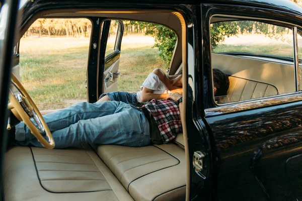 kissing people who are lying in the car and holding hands. look through the window in the car.side view.dressed in a plaid shirt, shorts and jeans, a white blouse. doors in the car are open, seats are spread out. black car with leather, white seats.d