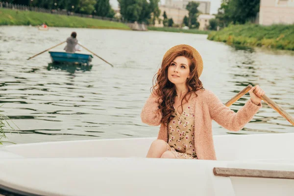 portrait woman is sitting in a white boat on the river. dressed in a flowered dress, a beige sweater, and a hat. summer vacation.in the distance one sees a retiring boat with a rower. date