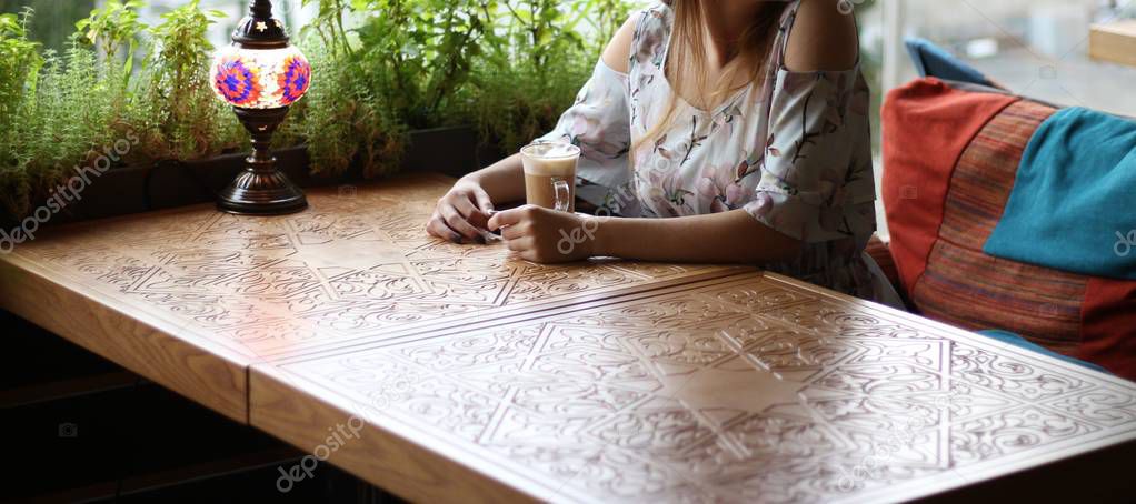 girl in a dress sitting at a table in a cafe and drinking coffee, waiting for a meeting, business, negotiations.dressed in a white dress with blue and pink flowers, sits by the window at the carved table, on which the lamp is burning.