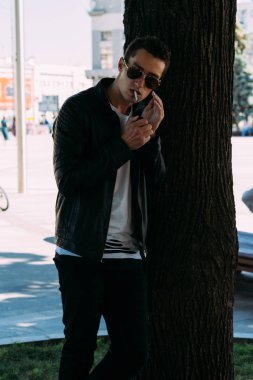 Genç bir adam bir ağaç kentin yakınındaki içiyor. Bir siyah ceket, kot pantolon, beyaz spor ayakkabı, beyaz bir T-shirt ve gözlük. kötü alışkanlık. halka açık bir yerde sigara