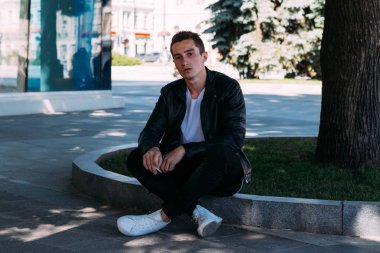 Genç bir adam bir ağaç kentin yakınındaki içiyor. Bir siyah ceket, kot pantolon, beyaz spor ayakkabı, beyaz bir T-shirt ve gözlük. kötü alışkanlık. halka açık bir yerde sigara