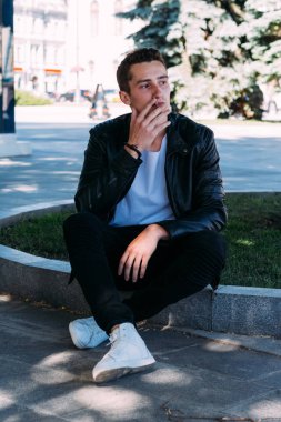 Genç bir adam bir ağaç kentin yakınındaki içiyor. Bir siyah ceket, kot pantolon, beyaz spor ayakkabı, beyaz bir T-shirt ve gözlük. kötü alışkanlık. halka açık bir yerde sigara