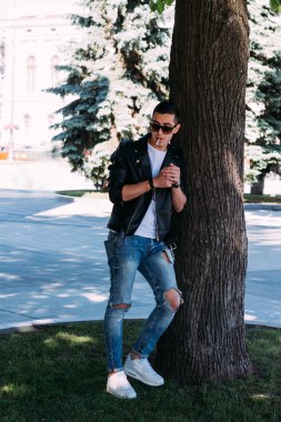 Genç bir adam bir ağaç kentin yakınındaki içiyor. Bir siyah ceket, kot pantolon, beyaz spor ayakkabı, beyaz bir T-shirt ve gözlük. kötü alışkanlık. halka açık bir yerde sigara