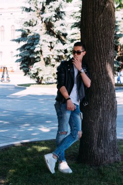 a young guy smokes near a tree in the city. A black jacket, jeans, white sneakers, a white T-shirt and glasses. bad habit. smoking in a public place clipart