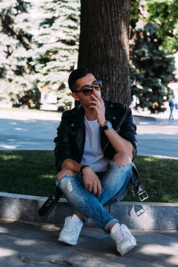 Genç bir adam bir ağaç kentin yakınındaki içiyor. Bir siyah ceket, kot pantolon, beyaz spor ayakkabı, beyaz bir T-shirt ve gözlük. kötü alışkanlık. halka açık bir yerde sigara