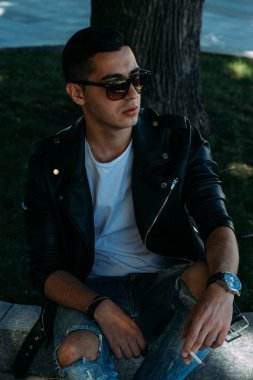 Genç bir adam bir ağaç kentin yakınındaki içiyor. Bir siyah ceket, kot pantolon, beyaz spor ayakkabı, beyaz bir T-shirt ve gözlük. kötü alışkanlık. halka açık bir yerde sigara