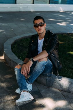 Genç bir adam bir ağaç kentin yakınındaki içiyor. Bir siyah ceket, kot pantolon, beyaz spor ayakkabı, beyaz bir T-shirt ve gözlük. kötü alışkanlık. halka açık bir yerde sigara