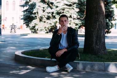 Genç bir adam bir ağaç kentin yakınındaki içiyor. Bir siyah ceket, kot pantolon, beyaz spor ayakkabı, beyaz bir T-shirt ve gözlük. kötü alışkanlık. halka açık bir yerde sigara