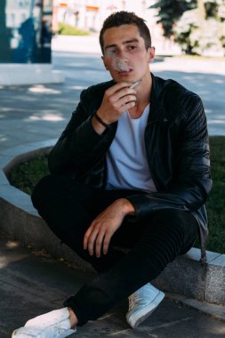 Genç bir adam bir ağaç kentin yakınındaki içiyor. Bir siyah ceket, kot pantolon, beyaz spor ayakkabı, beyaz bir T-shirt ve gözlük. kötü alışkanlık. halka açık bir yerde sigara