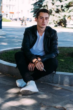 Genç bir adam bir ağaç kentin yakınındaki içiyor. Bir siyah ceket, kot pantolon, beyaz spor ayakkabı, beyaz bir T-shirt ve gözlük. kötü alışkanlık. halka açık bir yerde sigara