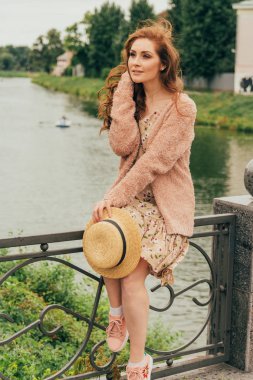 bir şapka, bir çiçek ve bej sweater.beautiful Kızıl saçlı girl.picture nehir, su, lake parkta, arka plan bej elbise elinde bir çit summer.horizontal fotoğraf oturur. romantizm, hayalperest