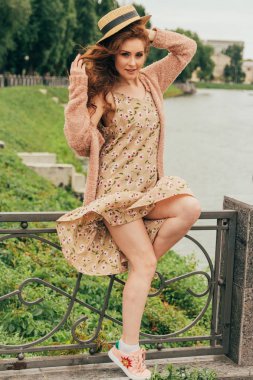 bir çit, şapkası, elinde bir çiçek ve bej sweater.beautiful Kızıl saçlı girl.picture nehir, su, Park, lake summer.the rüzgar pislikler kadar hem elbise ve saç alarak arka plan bej elbise oturur