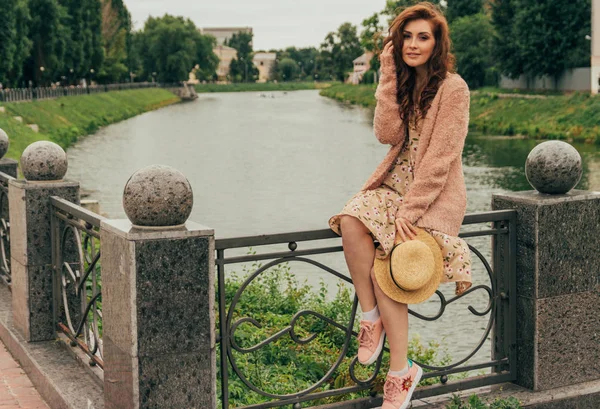 公園の湖の背景に美しい赤毛 Girl Picture 花とベージュの Sweater Sexuality とロマンス ベージュのドレス 帽子を手に フェンスの上に座っています — ストック写真