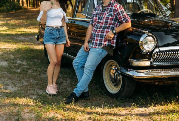 onun elinde bir çiçek bir kızla kot şort ve beyaz bir bluz, utanarak adam kot pantolon yaklaşımlar ve tarihleme için vintage ve retro bir araba üzerinde kırmızı bir gömlek bir kafeste duruyor. yaz