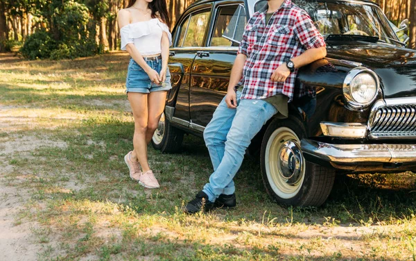 onun elinde bir çiçek bir kızla kot şort ve beyaz bir bluz, utanarak adam kot pantolon yaklaşımlar ve tarihleme için vintage ve retro bir araba üzerinde kırmızı bir gömlek bir kafeste duruyor. yaz