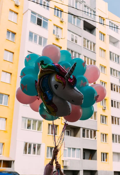 hands holding a bunch of colored balls of blue, green and pink with pink butterflies on them, an inflatable multi-colored unicorn, against the background of the building