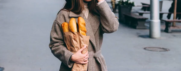 a girl in a coat and hat holds a bag with baguettes, bread, a loaf, on the street. a shopping trip,morning.