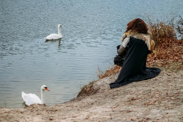 Medieval knight with sword in armor in the forest. autumn near the river.sits by the river on which the swan swims a man in armor, with a wolf cloak. costume games